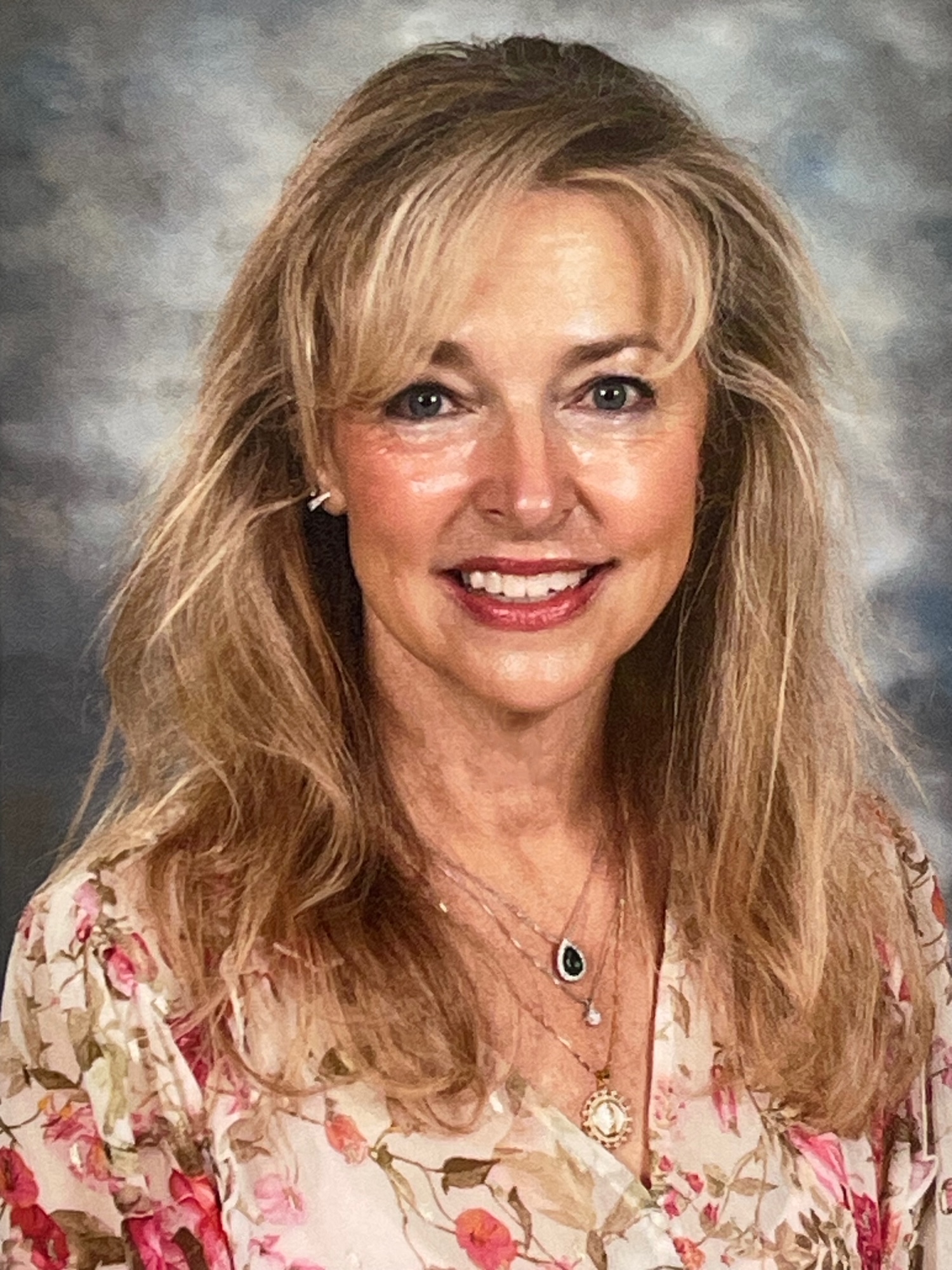 A woman with long, wavy hair smiles, wearing a floral blouse and layered necklaces, against a soft background.