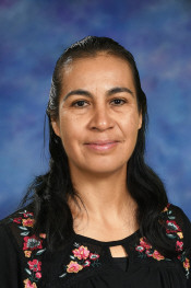 Smiling woman with long dark hair, wearing a black top with colorful floral embroidery, against a blue backdrop.