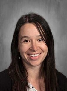 Smiling woman with long dark hair, wearing a black sweater, against a gray background.