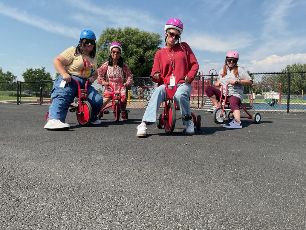 Five adults riding red tricycles in a playground, all wearing colorful helmets and enjoying the sunny day.