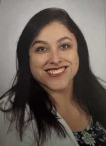 A smiling woman with long dark hair, wearing a floral top and a white jacket, against a light background.