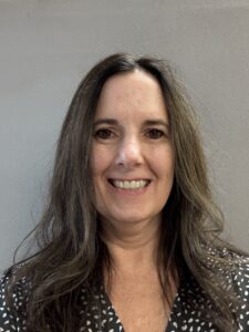 Smiling woman with long brown hair, wearing a patterned blouse, stands against a gray background.