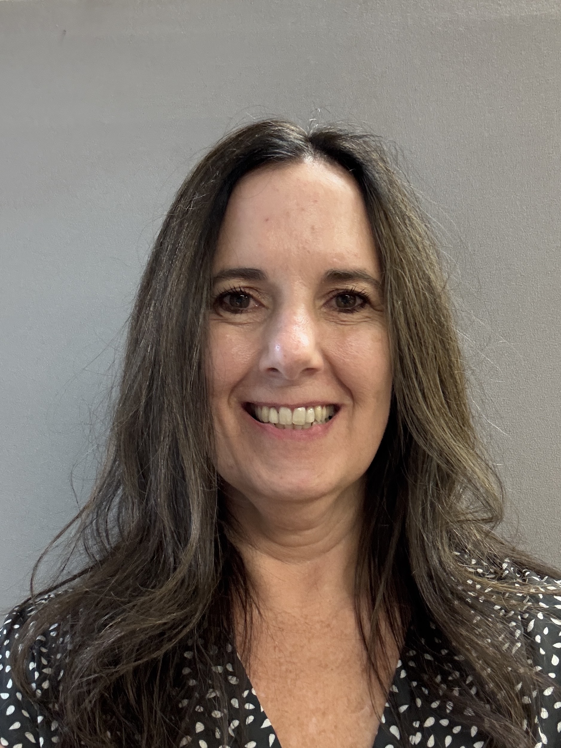 Smiling woman with long brown hair, wearing a patterned blouse, stands against a gray background.