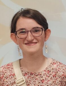 Young woman with glasses and floral shirt, smiling against a light background, wearing dangling earrings.
