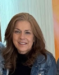 A smiling woman with long, wavy brown hair wearing a denim jacket, against a light-colored background.