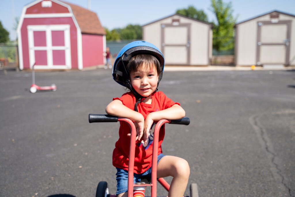 A young child in a red shirt and blue helmet smiles while sitting on a tricycle in an outdoor area.