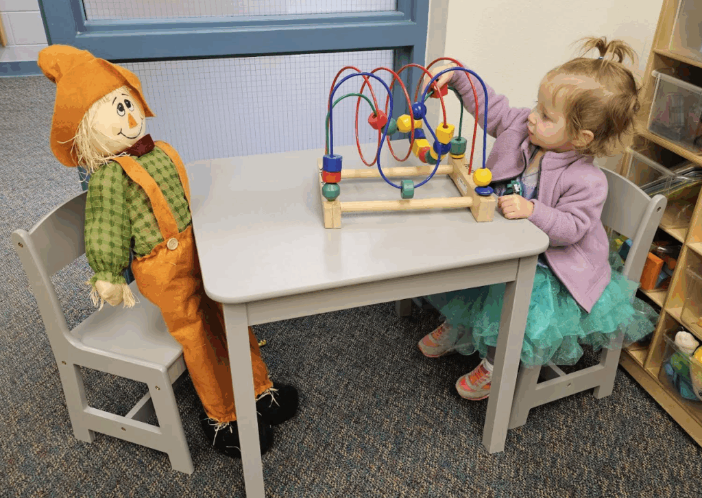 A child plays with a colorful bead maze at a table, next to a scarecrow decoration.