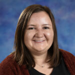 A smiling woman with shoulder-length brown hair, wearing a black top and maroon cardigan, against a blue backdrop.