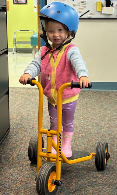 Toddler writing a scooter with a blue helmet, pink rain boots, and a pink vest.