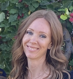 A woman with long, wavy brown hair smiles in front of green foliage with red and yellow accents.