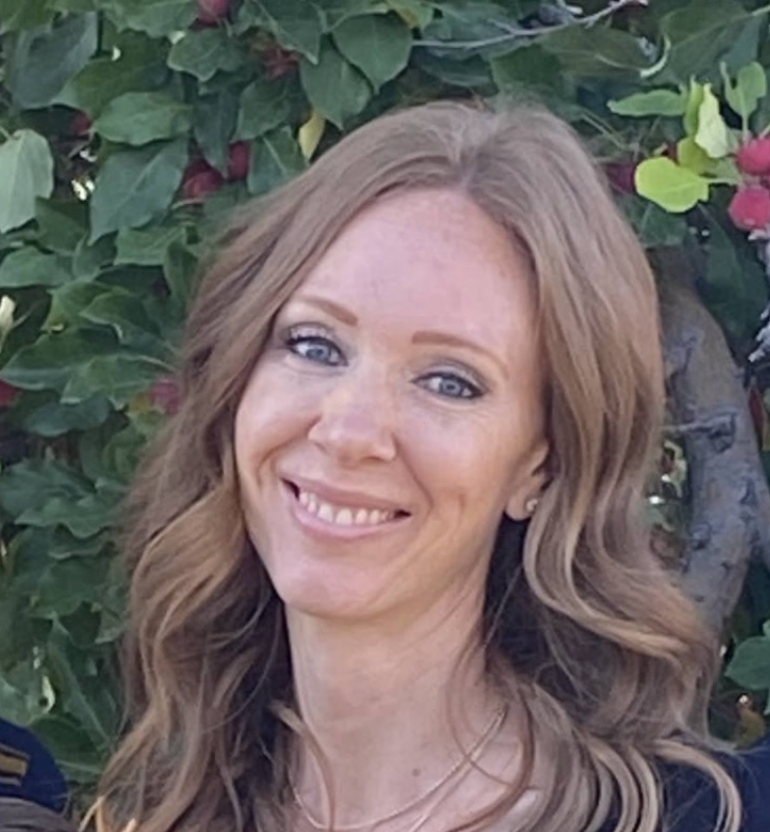 Smiling woman with wavy brown hair, wearing a black top, in front of greenery.