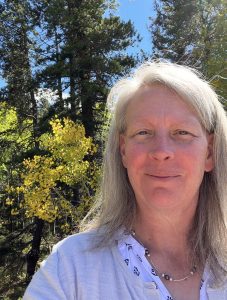 A person with long hair smiles in a forest with green trees and yellow leaves under a clear blue sky.