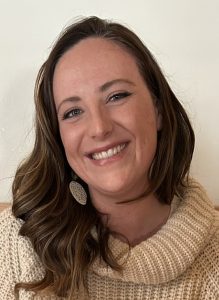 Mujer sonriente con el pelo largo, que lleva un jersey de punto y un pendiente grande, delante de un fondo liso.